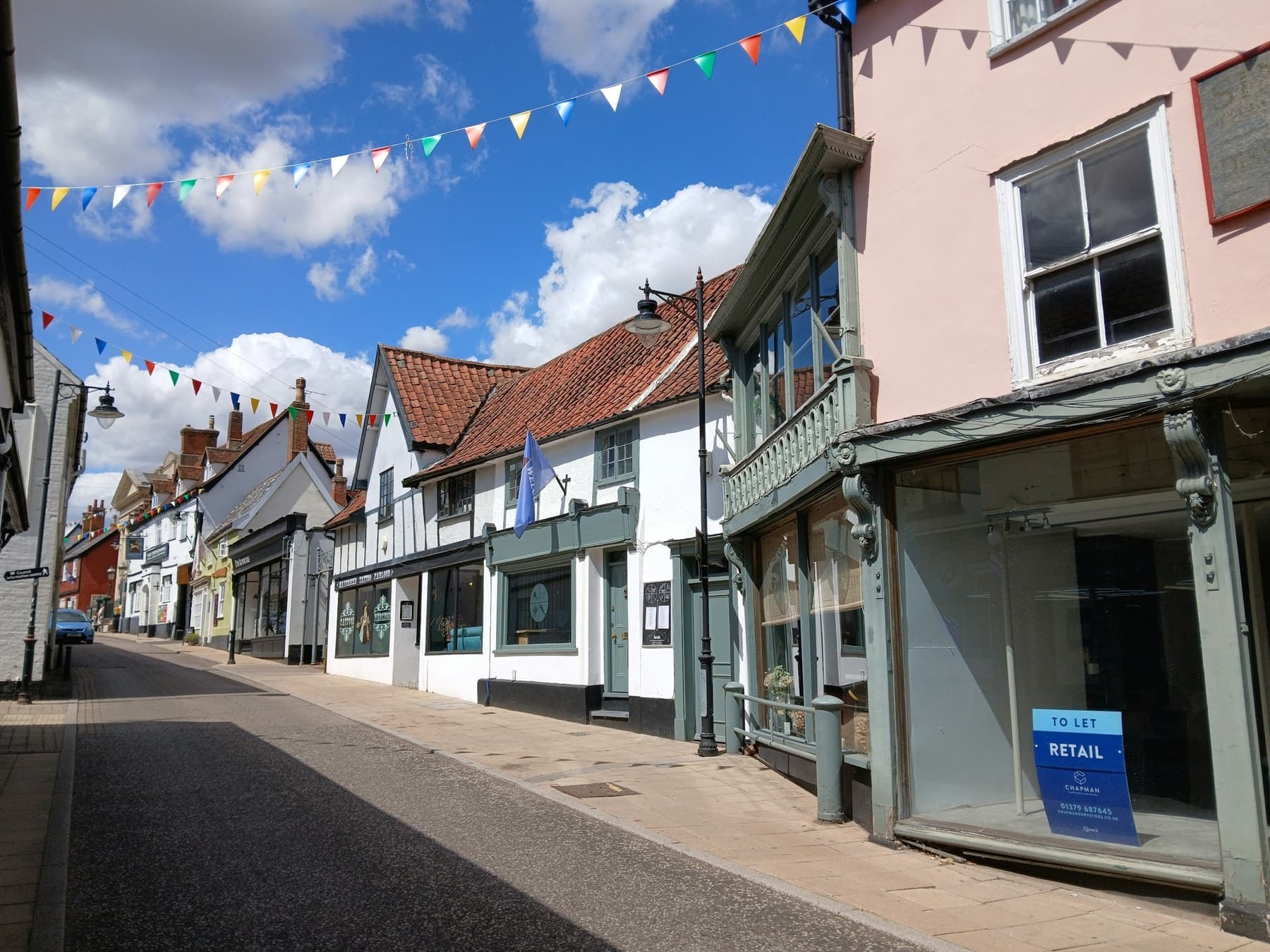 A sunny market-town high street with timber-framed and pastel shopfronts, and coloured bunting strung across a blue sky.